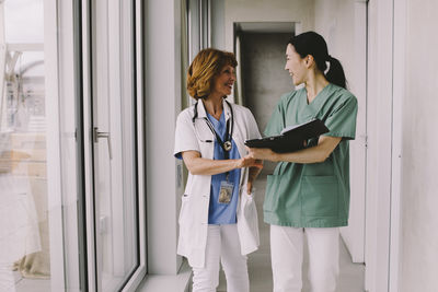 Smiling female medical colleagues discussing over record while walking in hospital corridor