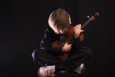 Boy playing violin while sitting against black background