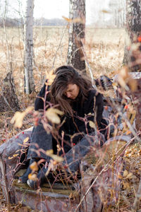 A young woman with long brown hair sits on the ground in a forest, eyes closed, smiling peacefully. 