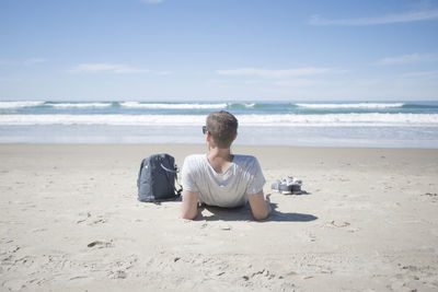 Rear view of man relaxing by backpack at beach on sunny day