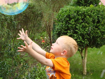 Boy reaching towards bubble by plants in yard