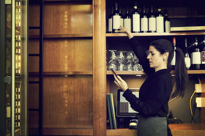 Side view of female owner checking list of wine bottles while standing in cafe