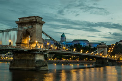 Illuminated bridge over river against cloudy sky