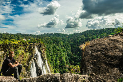 Portrait of man sitting on cliff against waterfall