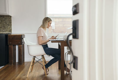 Full length side view of mid adult woman holding mobile phone while using laptop at home