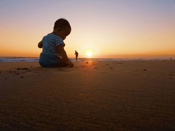 Rear view of baby boy sitting at beach against sky during sunset