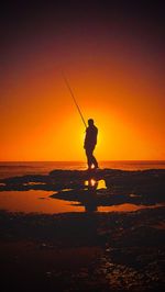 Silhouette man fishing in sea against orange sky