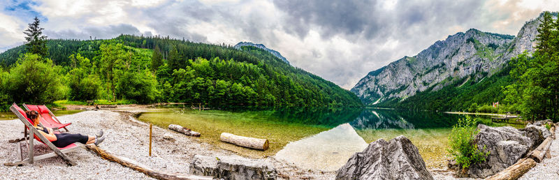 Panoramic view of woman sitting on lounge chair by lake and mountains against sky