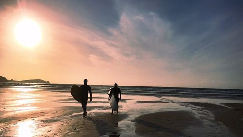 Rear view of surfers carrying surfboards while walking on beach during sunset