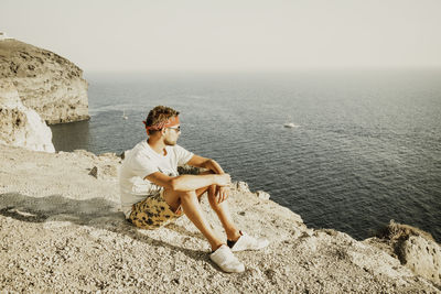 Man sitting on cliff by sea against clear sky