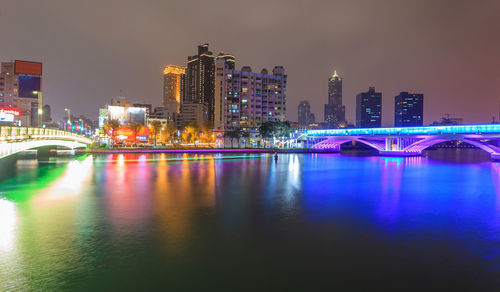 Illuminated bridge over river by buildings against sky at night