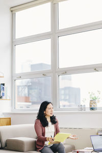 Businesswoman holding notepad while looking away against windows in office