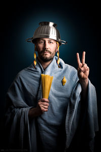 Portrait of man holding uncooked pasta while wearing colander on head against black background