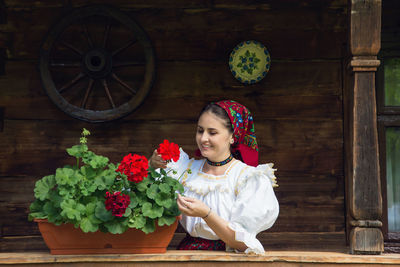 Happy young woman in romanian clothing looking at flowers on porch