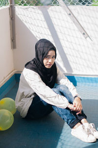 Portrait of young woman wearing hijab sitting on floor against wall