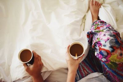 Low section of woman holding coffee cup on bed