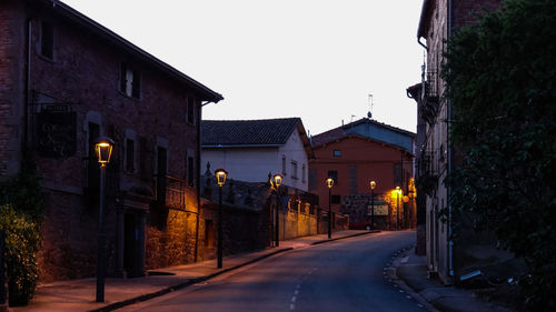 Empty road by illuminated lamp posts amidst buildings at dusk