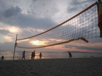 Volleyball net by people at beach against cloudy sky during sunset