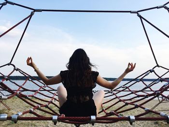 Rear view of young woman with arms outstretched sitting on hammock at lakeshore against sky