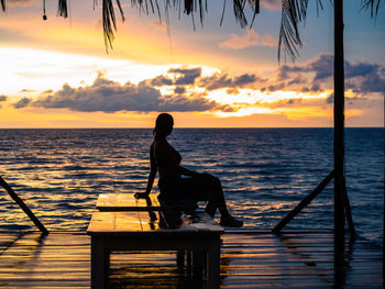 Silhouette woman sitting by sea against sky during sunset