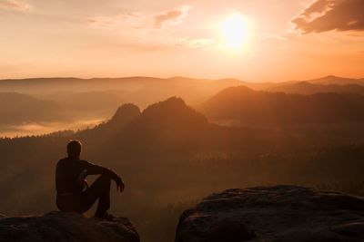 Man sitting on rock looking at mountains against sky during sunset