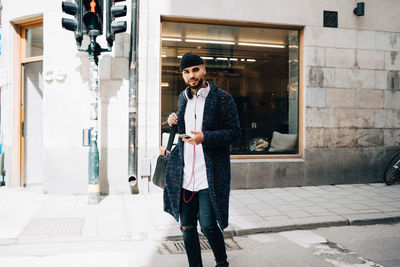 Portrait of confident young businessman walking with smart phone and bag against building in city