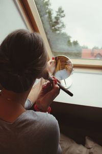 Rear view of young woman applying make-up against window at home