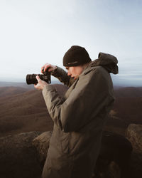 Side view of young man holding camera against sky