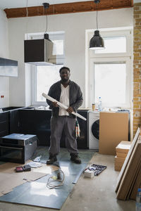 Full length of male carpenter holding blueprint standing in kitchen at apartment