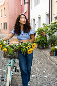 Beautiful young lady walking through the old city with turquoise bicycle and sunflowers in a basket.