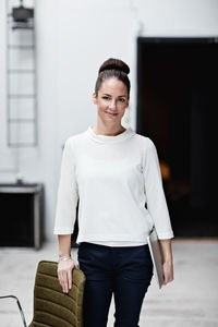 Portrait of smiling businesswoman holding digital tablet while standing by chair at office