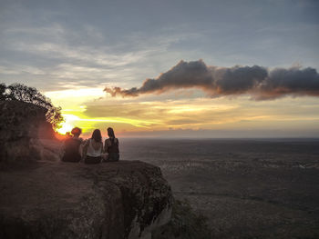 Rear view of friends sitting on cliff against cloudy sky during sunset