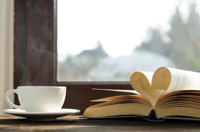 A cup of hot coffee with steaming next to heart shaped book on wooden table near by the window.