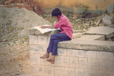 Boy studying while sitting at abandoned damaged retaining wall