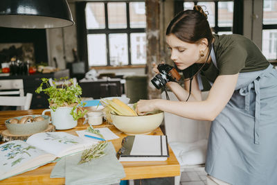 Female food stylist with digital camera setting up vegetable bowl on table in studio