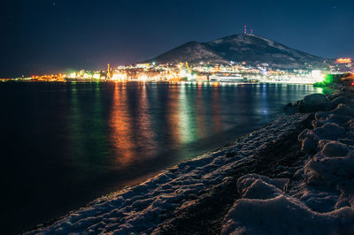 Illuminated city by sea against sky at night