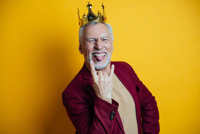 Portrait of smiling senior man wearing crown standing against yellow background