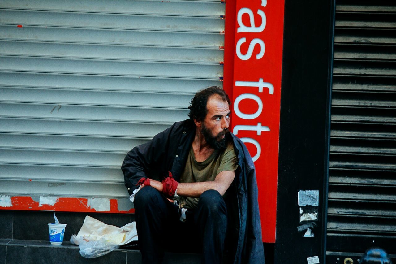 sitting, one person, three quarter length, lifestyles, real people, beard, casual clothing, front view, young men, facial hair, looking, men, day, communication, leisure activity, outdoors, wall - building feature, looking away, red, iron