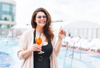 Stylish woman plus size body positive in sunglasses drinking with glass of cocktail near pool