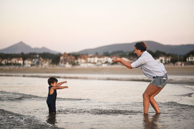 Side view of positive mother splashing water on cute son while standing together in shallow water of sea against residential buildings