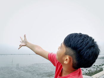 Close-up of boy with arms raised gesturing against sea and clear sky