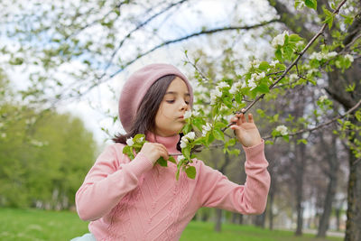 A little girl in a pink jumper holds a branch of a blossoming white apple tree in the park