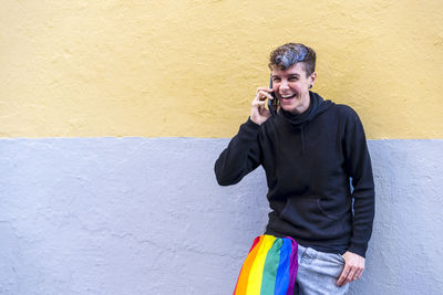 Non binary person with an lgbt rainbow flag talking on the phone outdoors.