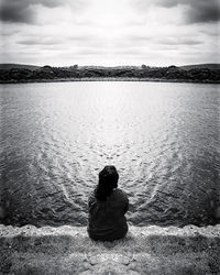 Rear view of woman sitting by lake against sky