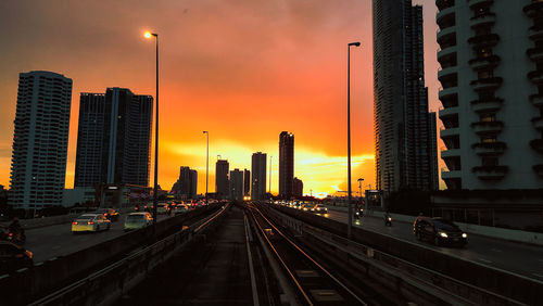 Vehicles on road by buildings against sky during sunset