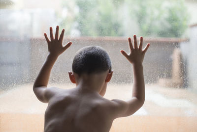 Rear view of shirtless boy looking through wet glass window