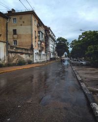 Wet road by houses in city against sky