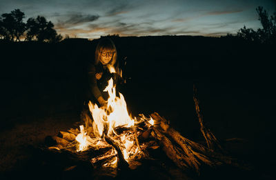 Woman sitting by bonfire at sunset