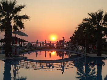 Man standing on footbridge over swimming pool against sky during sunset