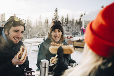 Happy friends eating hot dog while sitting at ski resort during winter